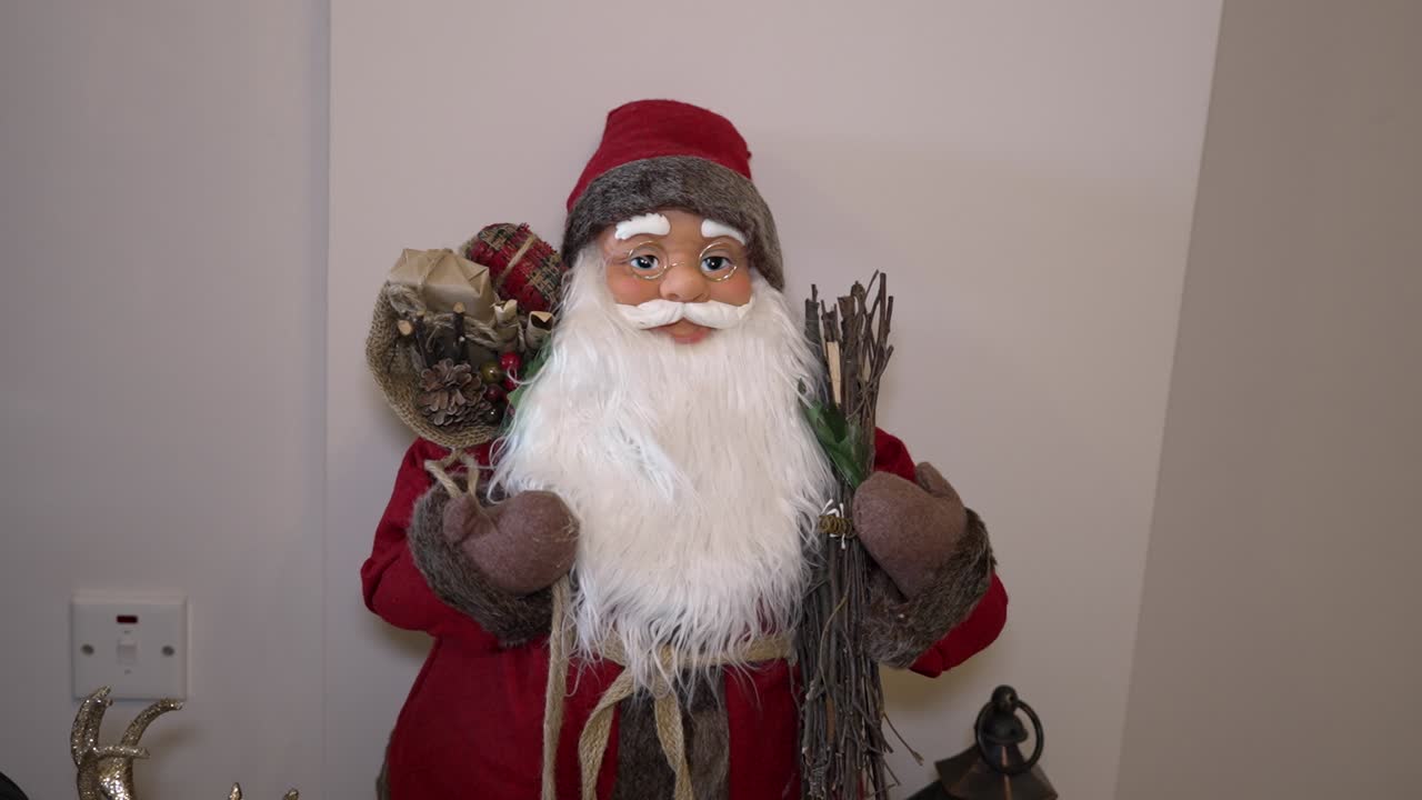 Se coloca una figura de Papá Noel en una sala de estar. Papá Noel viste de rojo y tiene barba blanca. Sostiene una bolsa y bastones. Decoraciones navideñas rodean la figura en el fondo.