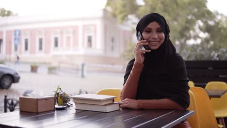 Joven-Y-Bonita-Mujer-Musulmana-Con-Hiyab-Hablando-Por-Teléfono-Y-Sonriendo,-Sentada-En-Un-Café,-Alegre