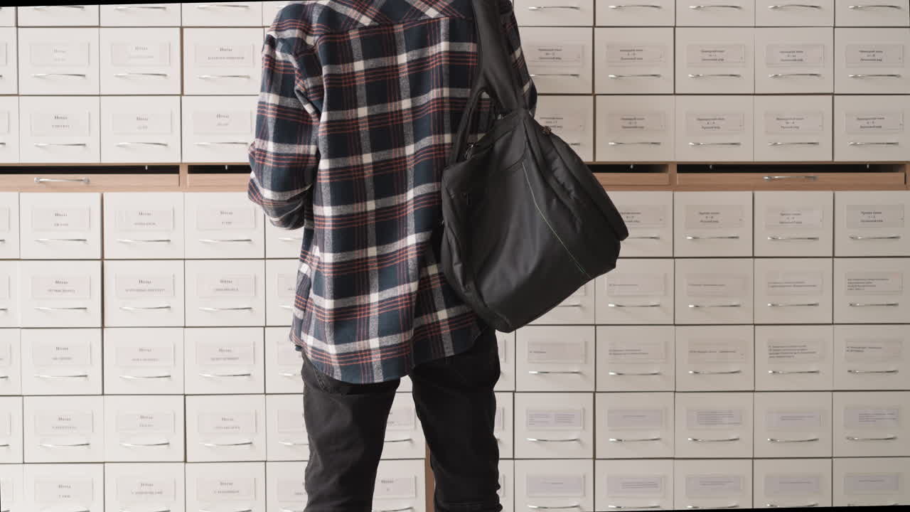 Man stands in front of card index boxes in library achieve. Student in checkered shirt with backpack seeks information in drawer unit. Guy in paper storage
