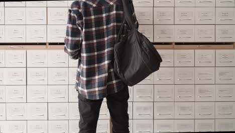 man stands in front of card index boxes in library achieve. student in checkered shirt with backpack seeks information in drawer unit. guy in paper storage