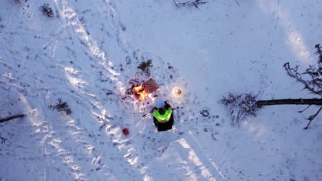 man sitting on a chair next to a fire and lamp in a forest covered with snow