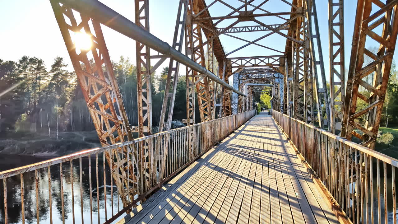 Sunlight flares through the steel trusses of a bridge with a wooden pedestrian deck