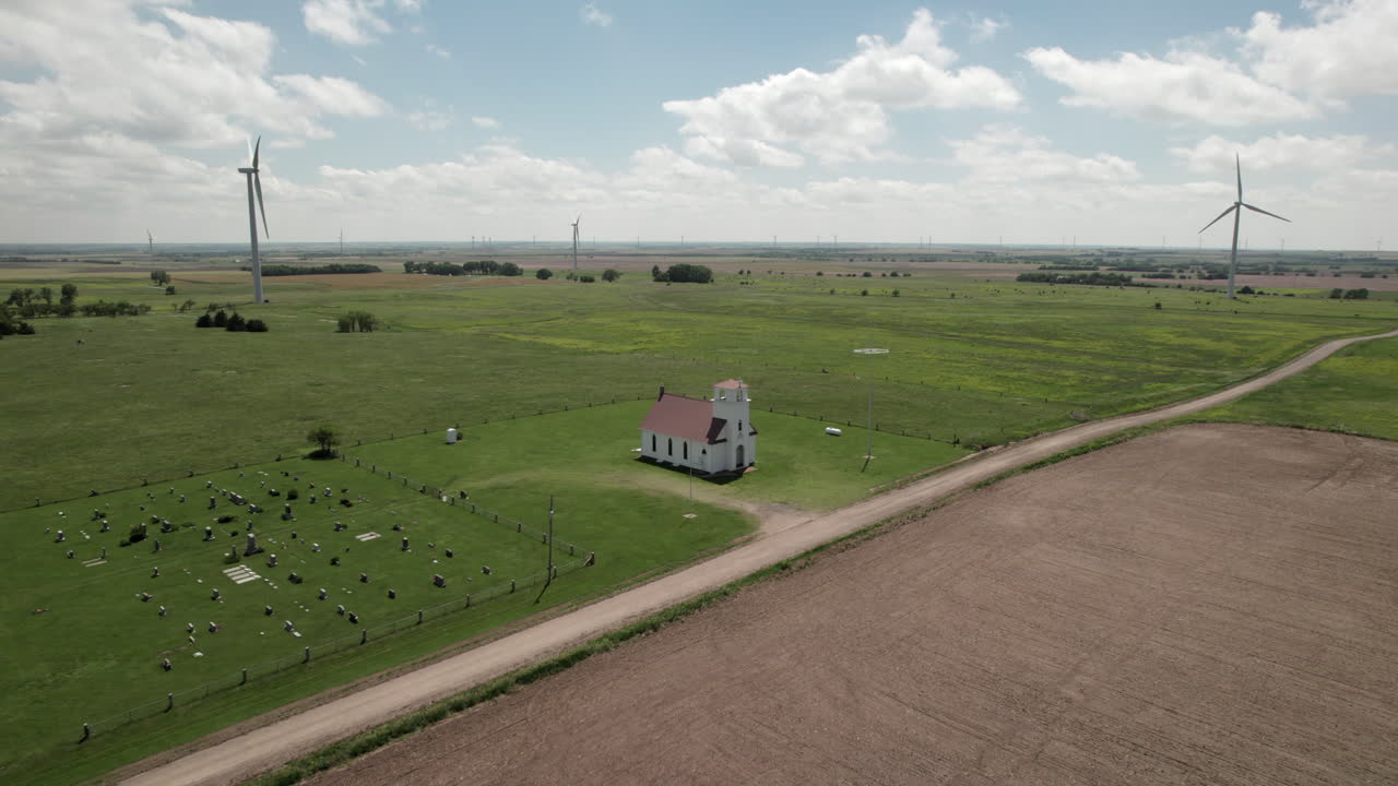 Slow aerial approach towards small church, cemetery. Wind turbines in background.