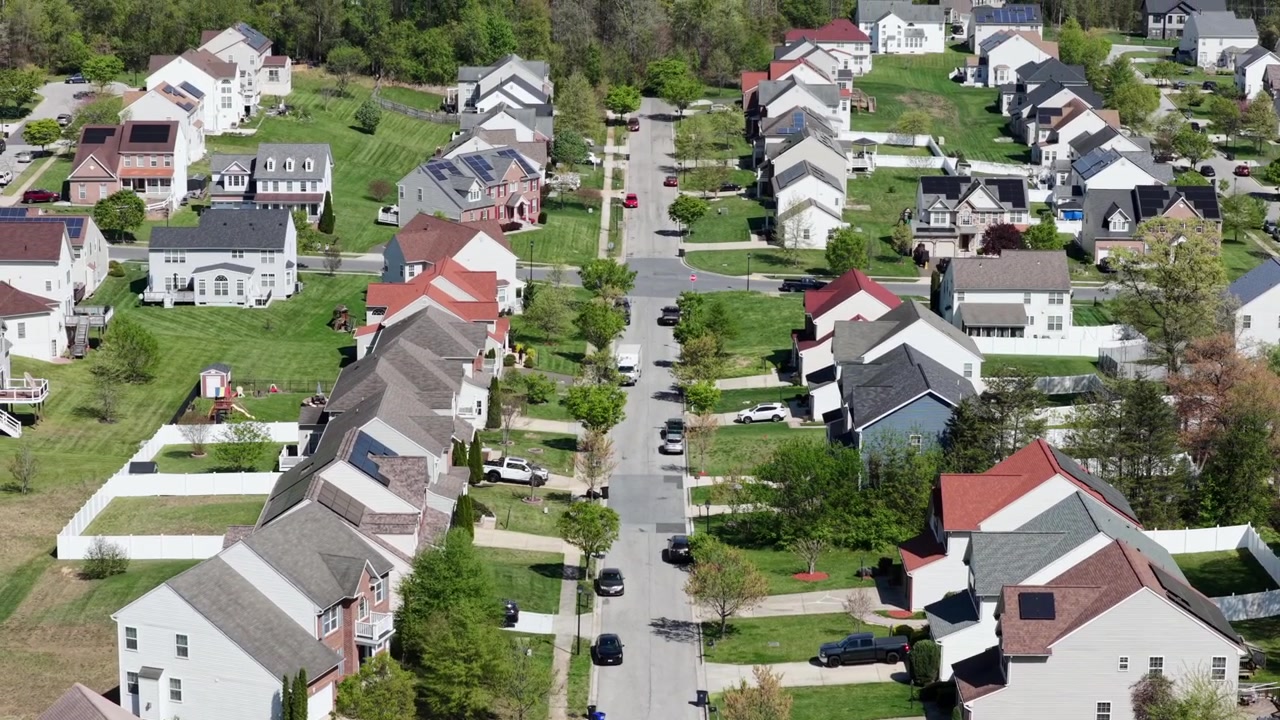 Quartiere residenziale americano con case unifamiliari, auto parcheggiate lungo la strada e giardini verdi in primavera sotto un cielo sereno che mostra una tranquilla comunità residenziale. Vista aerea da drone negli USA