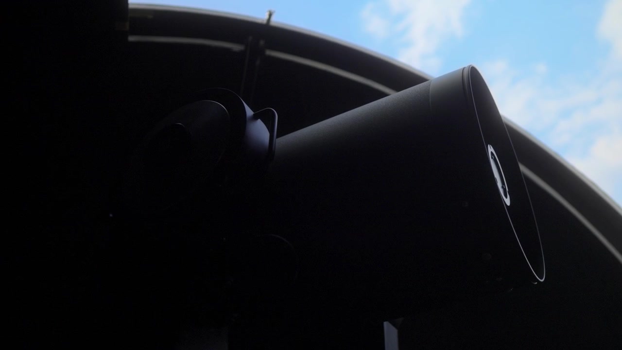 Silhouette scura di un grande telescopio astronomico posizionato all'interno di una cupola di osservatorio, che guarda attraverso il tetto aperto verso un cielo blu con nuvole