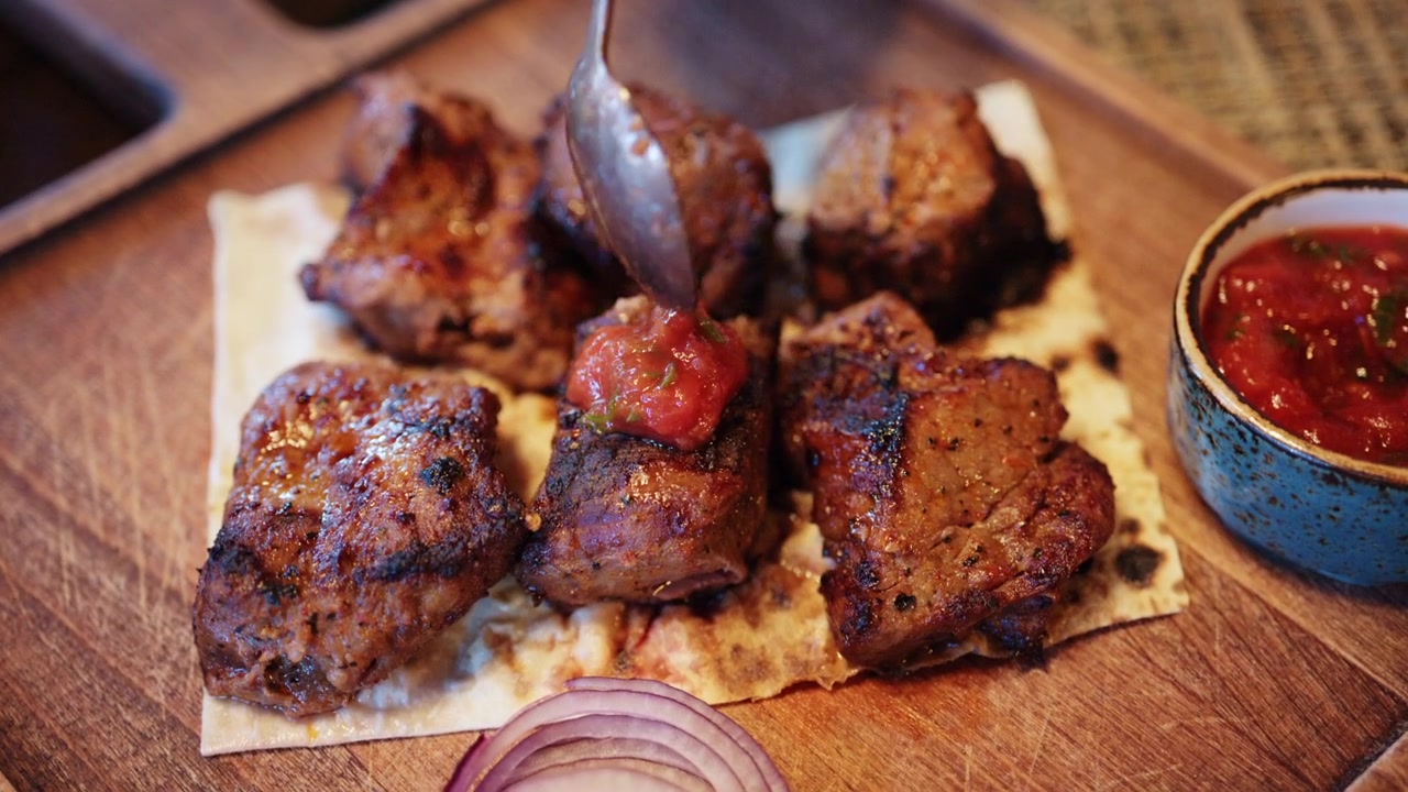 Lähikuva grillatuista lihapaloista aseteltuna leivän päälle, samalla kun henkilö leikkaa yhtä palaa veitsellä ja haarukalla.
