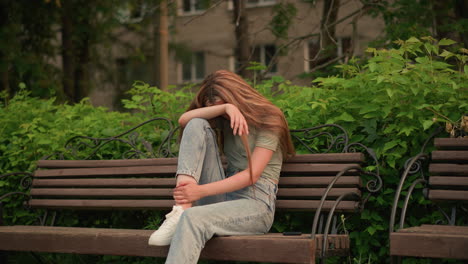 lady in casual jeans and white sneakers sits on wooden bench in park, resting head on arm in reflective, somber posture with hair cascading down her shoulder, surrounded by lush greenery and trees