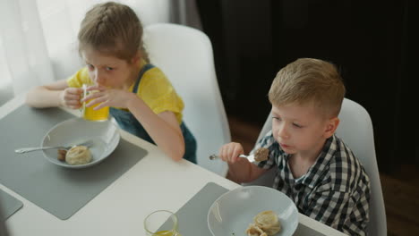 overhead view of young boy eating food while girl in yellow top drinks juice, seated at white dining table with bowls and placemats, bright window light