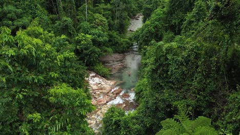 drone shot panning up from the colorful jungle, revealing the tiny waterfall and its streams over the rocks, creating a small river where a young man is standing with his arms reaching for the sky