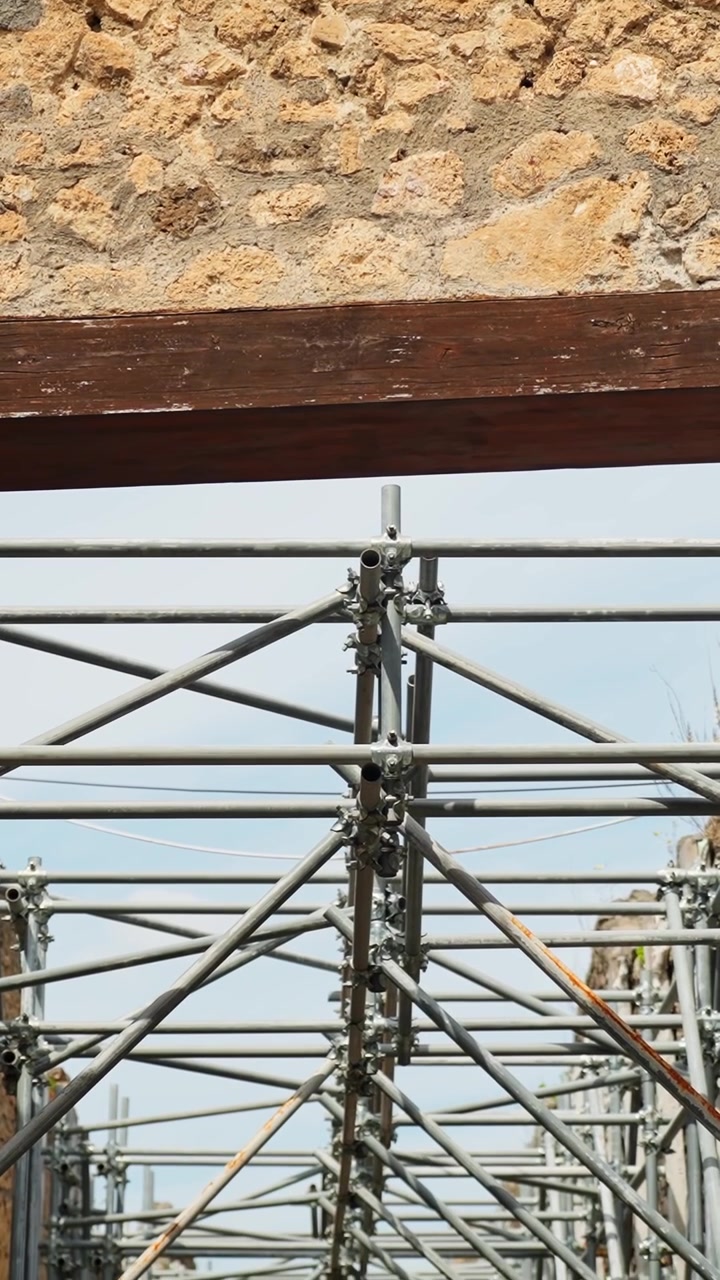 Vertical view of metal scaffolding supporting ancient Pompeii masonry, with rough stone wall texture, wooden beam, blue sky, and restoration structure details.