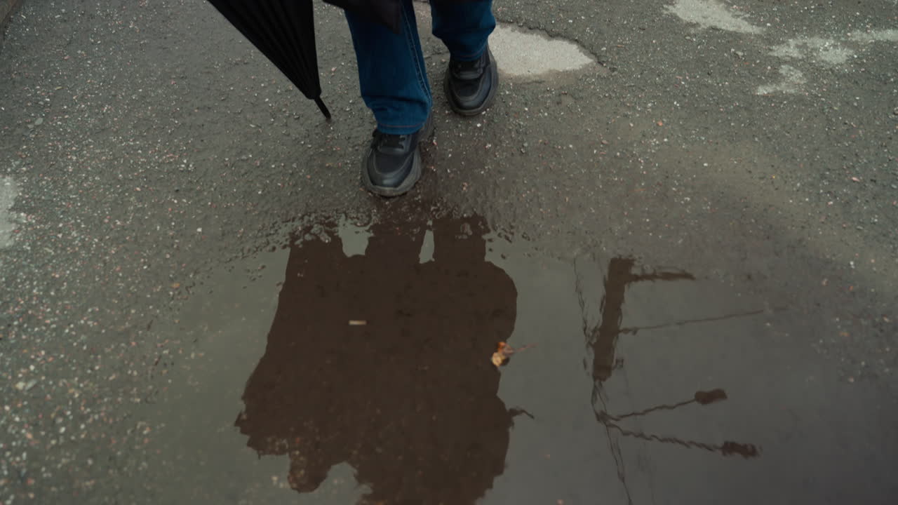 Donna bianca in piedi sui binari del tram che tiene un ombrello chiuso, indossa jeans di denim e stivali; attraversamento urbano con rotaie e riflesso di pozzanghera, asfalto bagnato increspato, pausa contemplativa, umore da pendolare, città sottile