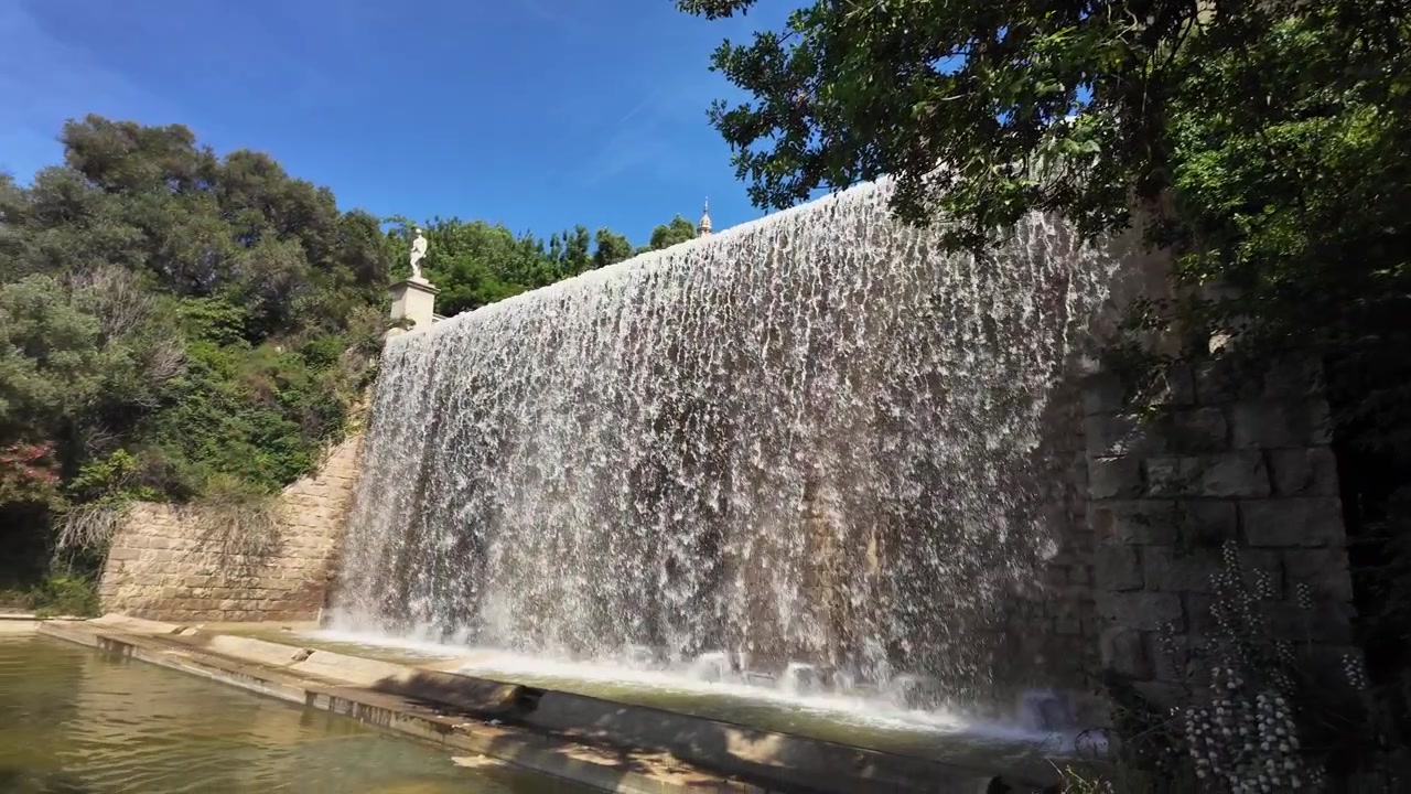 Riprese dinamiche a mano libera catturano l'iconica fontana a cascata di Montjuic al rallentatore, con la luce del sole che luccica sull'acqua impetuosa, circondata da una vegetazione lussureggiante e dal cielo azzurro e limpido di Barcellona.