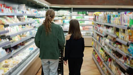 Teen-girl-and-her-mom-shopping-in-the-supermarket-with-cart,-rear-view