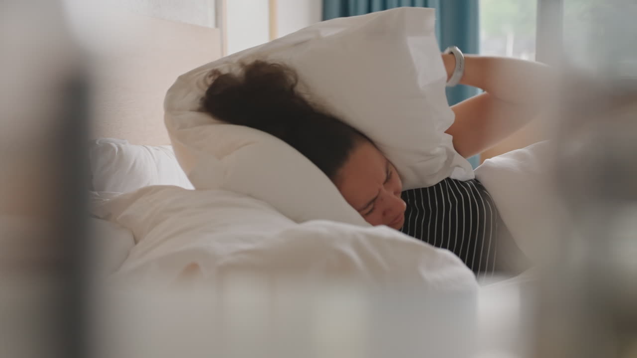 Udmattet hotelgæst dæmper lyd med en pude, mens hun ligger ved vinduet. Et sidebillede gennem en sløret forgrund afslører en kvinde i prikket tøj, der dækker ørerne med en blød pude, omgivet af turkise gardiner og et natbord.