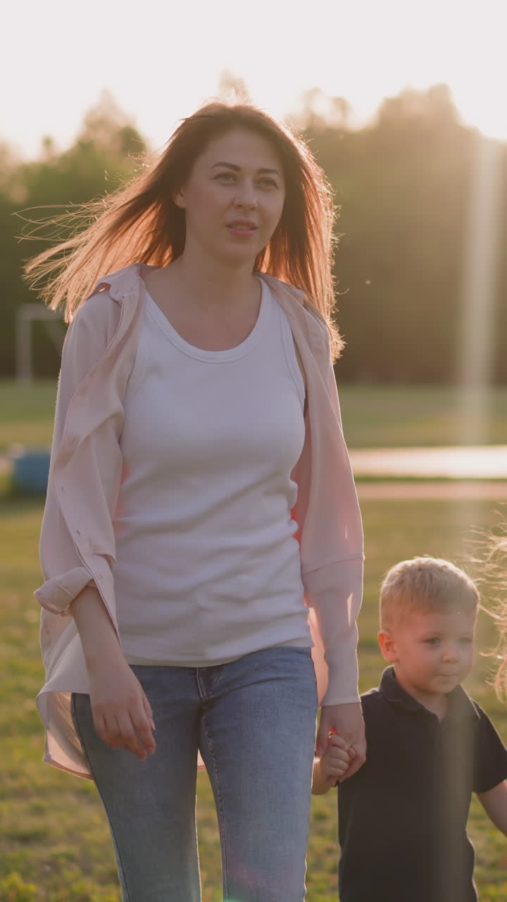 Happy woman with little kids walks along city park in summer evening slow motion. Mother with cute children at promenade. Rest and enjoyment of weather