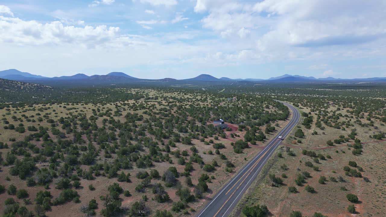 Ripresa aerea ascendente che mostra una strada a due corsie a Williams, in Arizona, che curva dalla parte inferiore sinistra attraverso il centro dell'inquadratura e poi di nuovo in lontananza. Ci sono montagne e cielo azzurro punteggiato di
