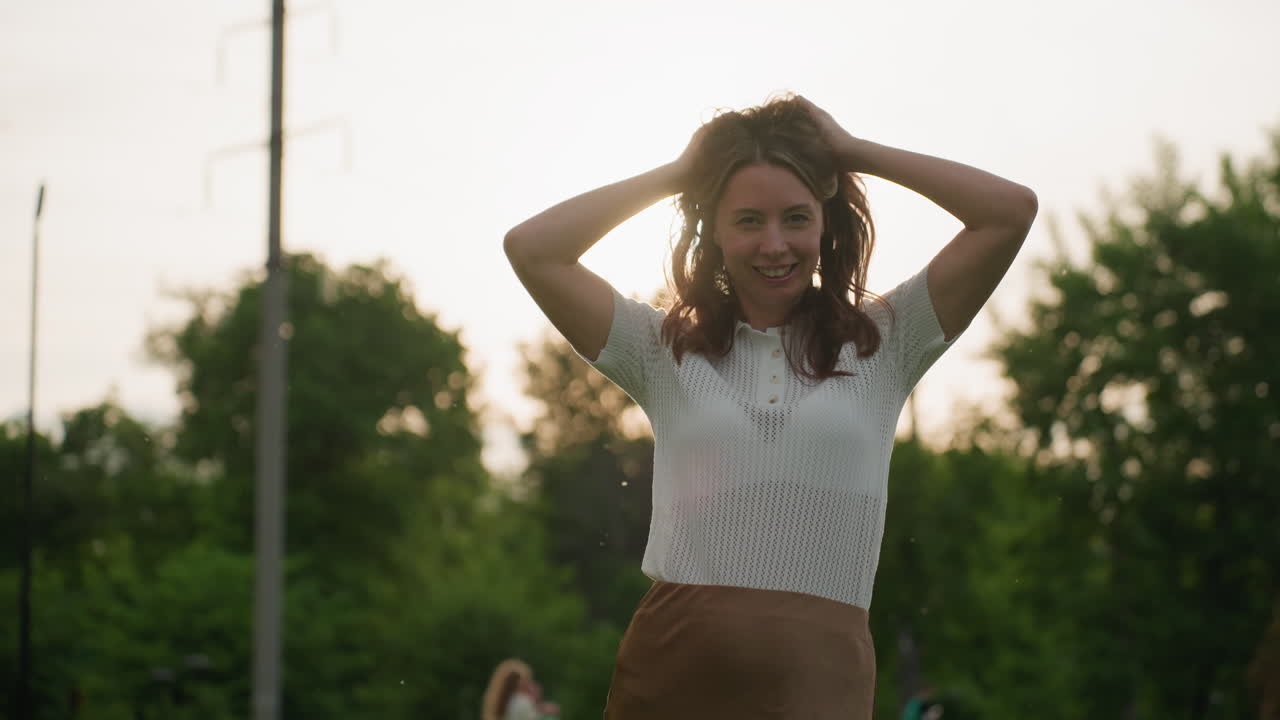 medium shot beautiful lady playing with hair while smiling outdoors, sun backlight creating warm glow, green blurred background, carefree joyful expression during relaxed park outing