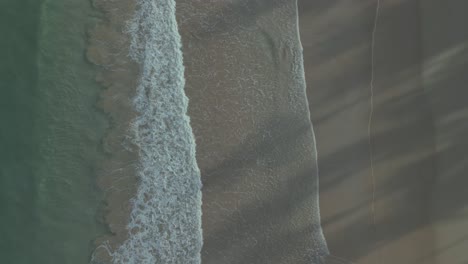 Ocean-waves-calmly-wash-over-sand-beach,-top-down-aerial-pull-out