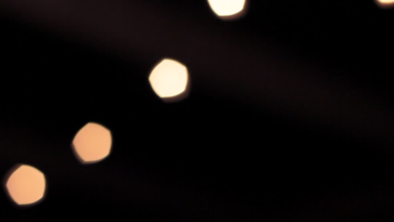 Golden orbs blur into view, sharpening into glowing string lights as the camera pans right, revealing their warm brilliance against the night