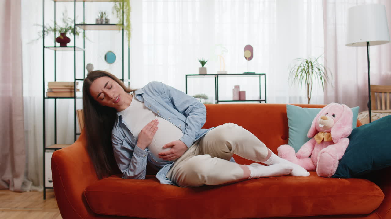불안한 젊은 임신 한 여성은 아프고 복통으로 고통 받고 집 소파에 누워 있습니다.