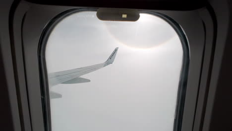 Smooth-push-in-between-airplane-window-to-view-of-wing-tip-against-clouds,-sunburst-around-sun