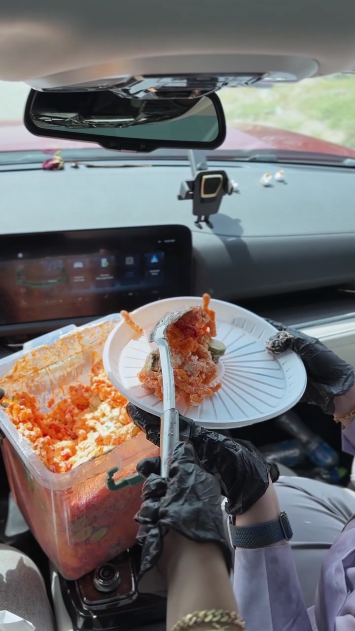 Close up of a person wearing black gloves serving a messy orange snack salad from a plastic container to a plate inside a modern car during a road trip.