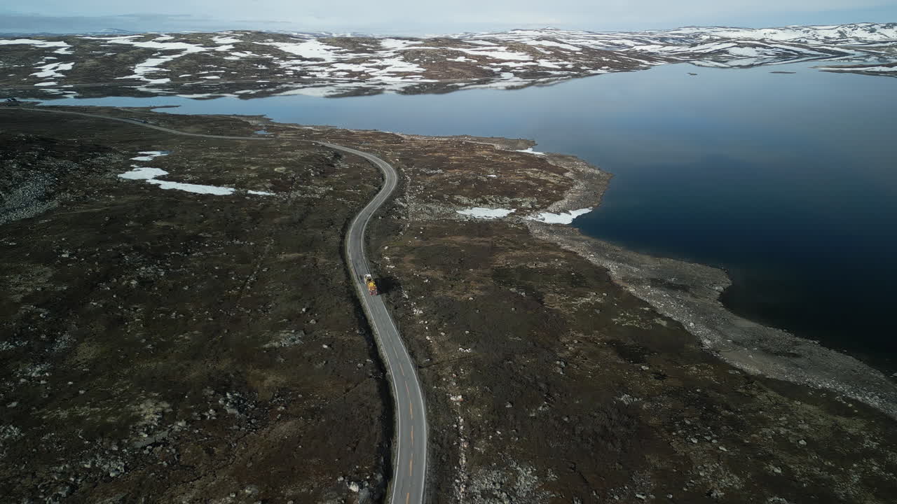 Drone shot of a truck driving on the road through Hardangervidda Park. The road runs along a lake near Haugestøl in Norway. Snow-covered mountains and the sky are reflected in the flat, calm water