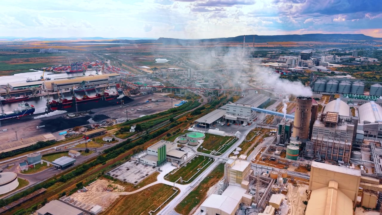 Industrial factory beside cargo ships and railway tracks. Steam rises over processing halls while cranes and vessels line the waterfront terminal.