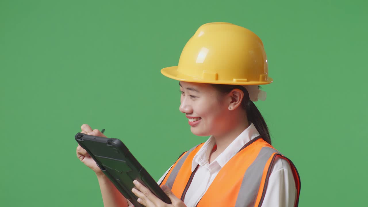안전 헬을 입은 아시아 여성 엔지니어가 녹색 스크린 배경 스튜디오에 서 있는 동안 태블릿에 메모를 하고 주변을 둘러보고 있습니다.