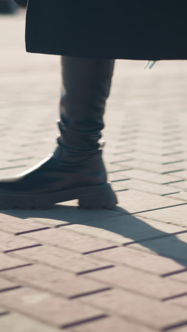 vista de la pierna de una persona con elegantes botas de cuero y abrigo chic caminando con confianza en el pavimento entrelazado bajo la luz del sol, sombra en el suelo, características de fondo borrosas coches estacionados
