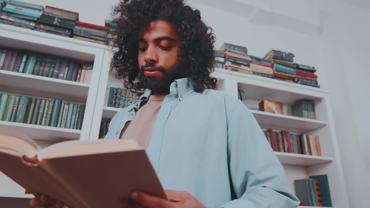 Young focused middle eastern man reads and flips through book stands in library
