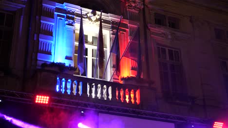 a french town hall decorated at night for a show, with blue, white, and red lights and a french flag