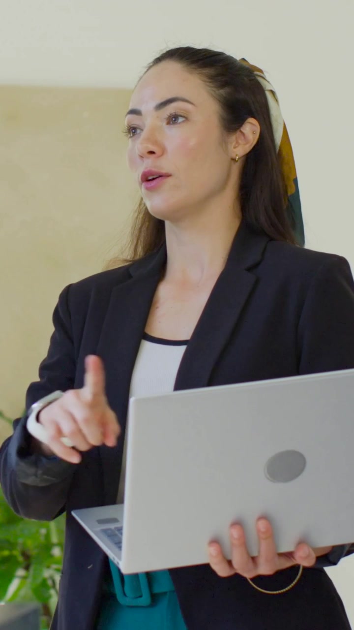 Vertical video: Glancing at silver laptop, woman in blazer adjusting grip and presenting at office