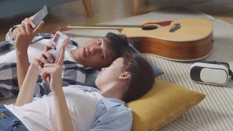 asian teen couple playing smartphone while lying on carpet on the floor at home. laughing, speaking, and enjoying time together