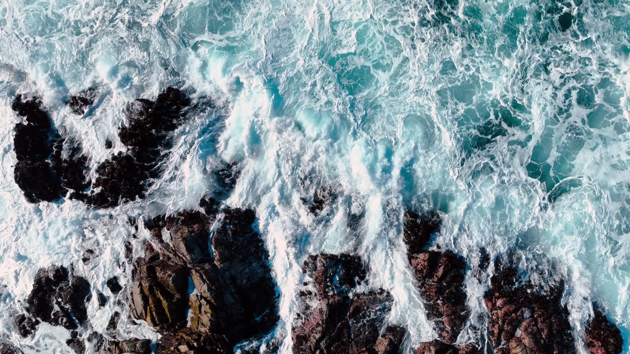 Abstraktní letecká textura oceánského příboje narážejícího na skalnaté pobřeží. Záběr z vysokého úhlu bílé vody a mořské pěny vířící kolem tmavých pobřežních kamenů