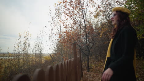 side view of young lady in yellow beret and black coat, walking up to decorative wooden fence, resting her hand, gazing up at autumn trees with dry leaves scattered around, river in background