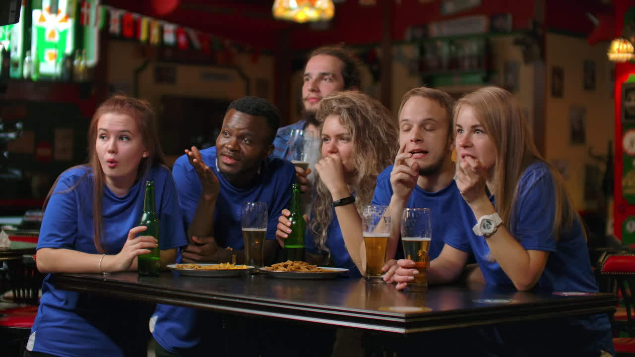A multi-ethnic group of friends of fans in blue t-shirts will watch a match on TV in a bar watch a tense exciting moment and celebrate a goal scored. Hugging having fun and making a glass of beer.