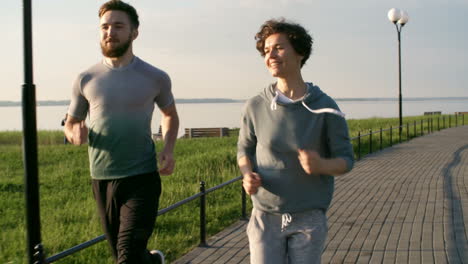 Sporty-Young-Man-And-Woman-Smiling-And-Talking-While-Running-Together-On-Riverside-Promenade-In-The-Morning