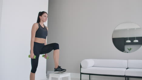 A-young-brunette-woman-performs-concentrated-pacing-on-a-chair-in-a-bright-interior-apartment-with-dumbbells-in-her-hands.