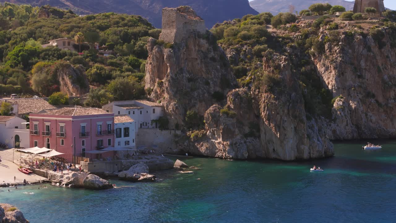 Aerial drone shot lowering down revealing a resort and private beach with huge rocks at Tonnara di Scopello, Sicily, Italy, showcasing the coastline during Italian summer