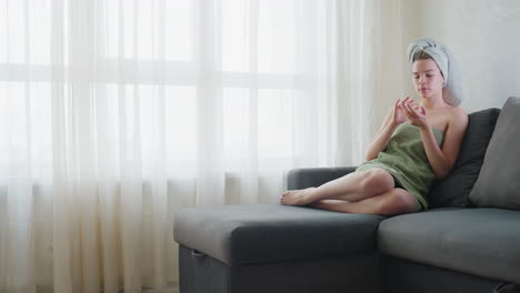 lady wrapped in green towel sits with legs crossed on couch near bright window, gently applying liquid skincare product to forehead with focus and care in peaceful indoor setting