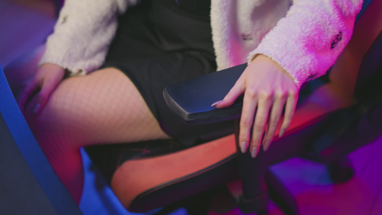 Aerial view of woman seated in gaming chair adjusting armrest with one hand while other hand rests on leg, dressed in fluffy sweater and fishnet tights under purple and pink lighting in tech space