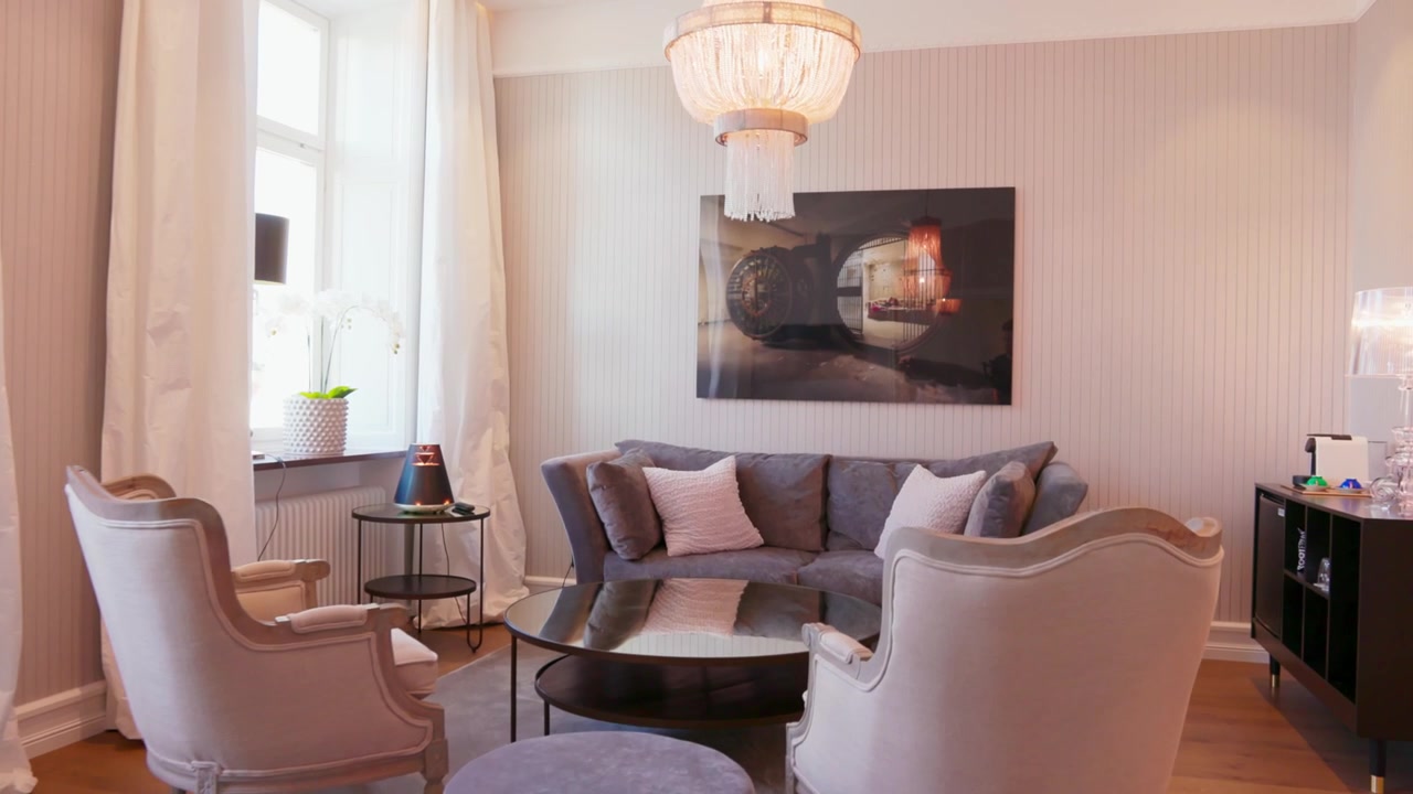 Brede gimbalopname van een verfijnde woonruimte met pluche zitmeubelen, stijlvolle kroonluchter, transparante witte gordijnen, grote ramen met natuurlijk licht, houten vloeren die klassieke en eigentijdse elegantie combineren