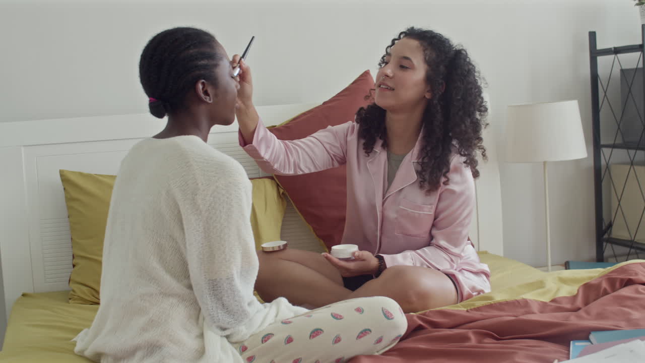 Girl Doing Makeup to Friend Sitting on Bed