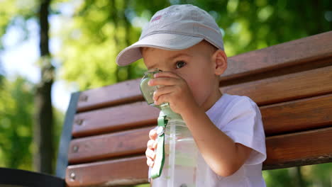 lovely kid in cap opening the bottle. cute baby boy drinking water on the bench in the park. low angle close up.