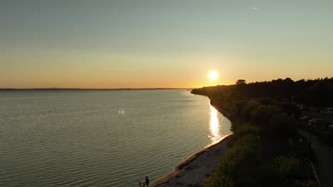 Aerial-view-of-a-sunset-over-a-calm-body-of-water-with-a-beach-and-two-people-walking-along-the-shore-in-Kuźnica,-Poland