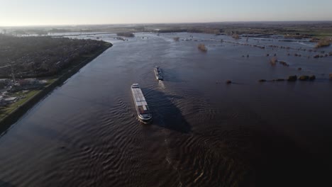 aerial following of inland shipping large cargo vessels leaving ripple waves on river ijssel during high water level with flooded floodplains of tower town zutphen in the netherlands