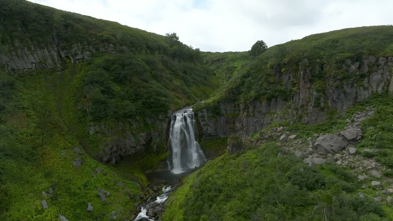 Riprese drone di una potente cascata che si getta in una gola fluviale rocciosa circondata da rigogliose colline verdi e scogliere nel Territorio della Kamchatka, Russia, in una giornata estiva nuvolosa