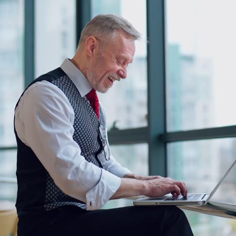 happy senior man typing on laptop near the big window. excited freelancer working on a computer at home. remote freelance work on retirement. active modern lifestyle of older people.
