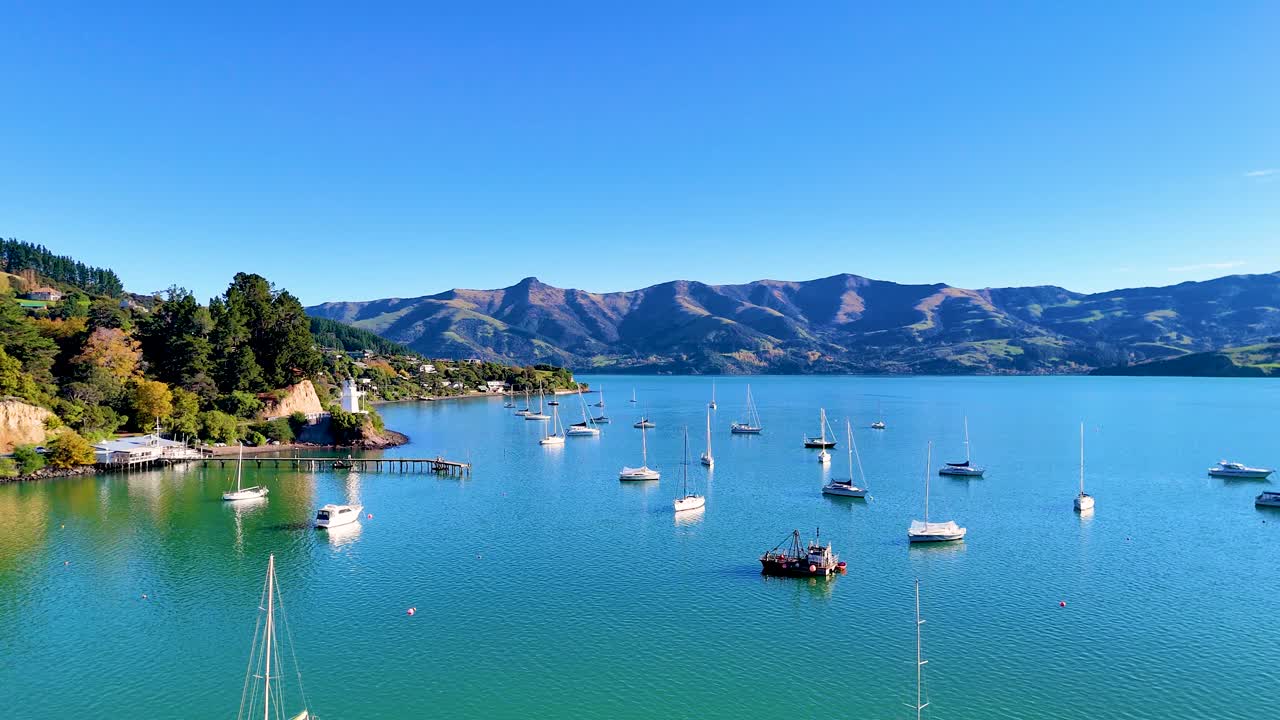 Boats gently float in Akaroa Harbour under clear blue skies, surrounded by lush hills and calm waters